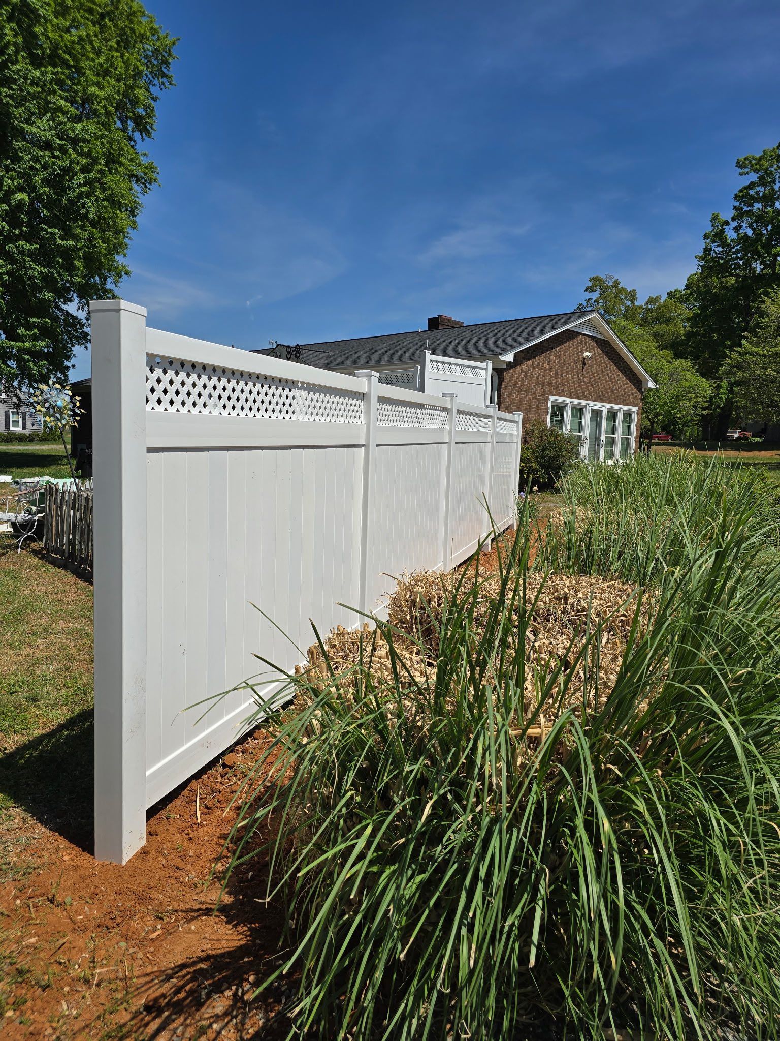White vinyl fence beside a garden bed with tall grasses, against a blue sky and brick house.