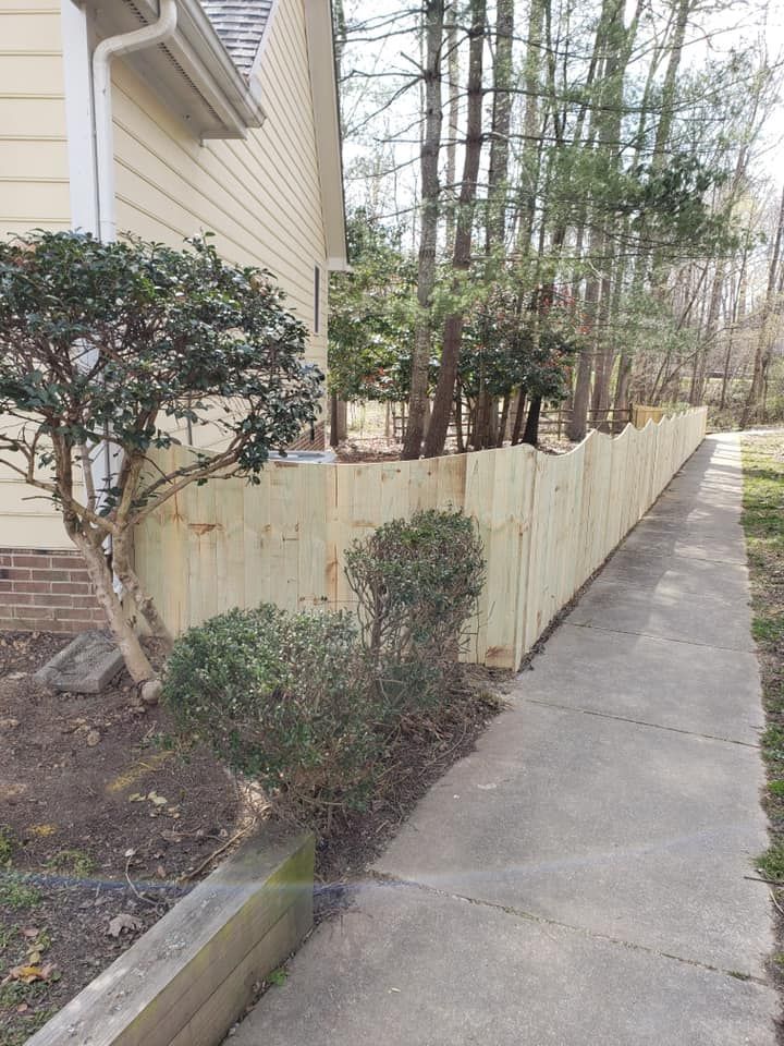 Sidewalk beside a beige house with a wooden fence and shrubs along a narrow garden bed