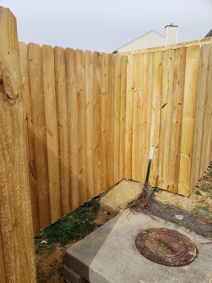 Wooden fence corner beside a concrete path with a metal manhole cover and shovel leaning nearby
