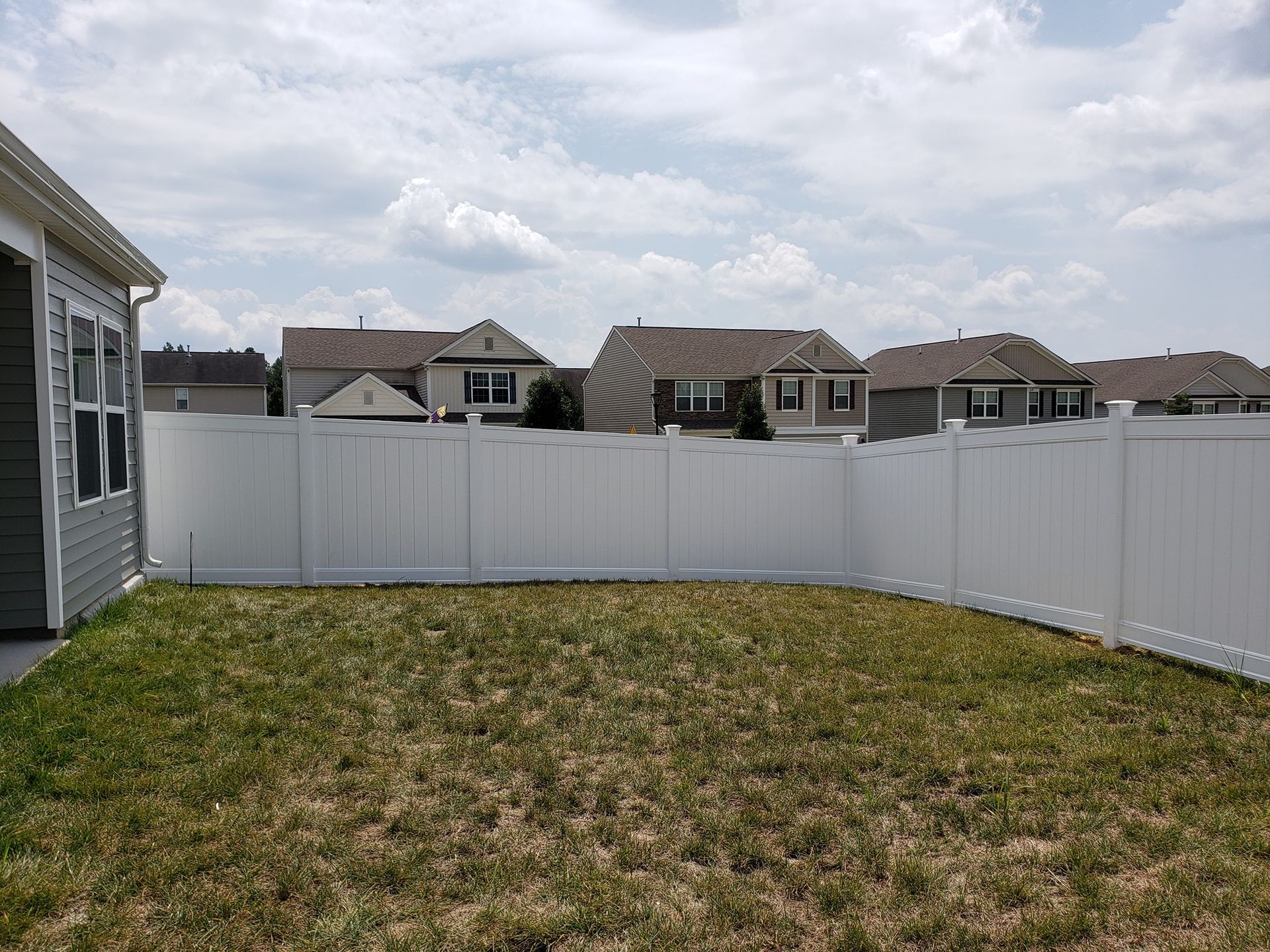 Small fenced backyard with patchy grass, a gray house on the left, and cloudy sky overhead.