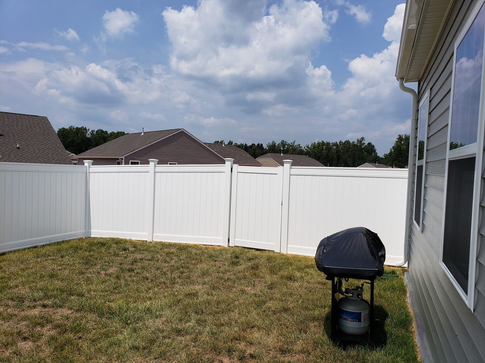 Backyard with white vinyl fence, patchy grass, grill, and cloudy sky beside a house