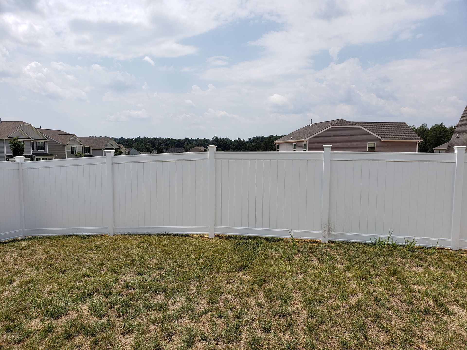 White vinyl fence across a grassy backyard under a cloudy sky, with houses visible beyond.