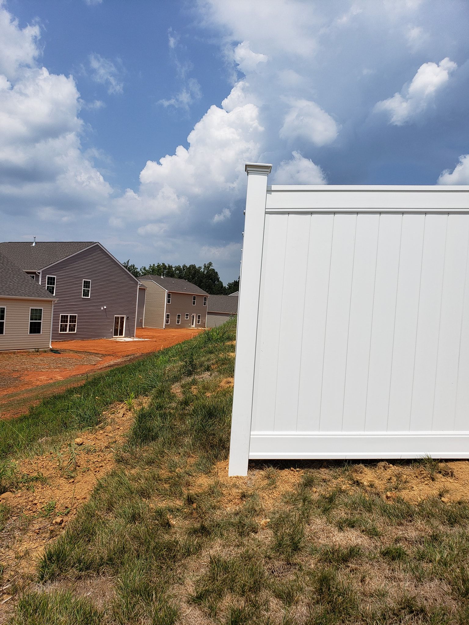 Suburban construction site with white vinyl fence, dirt path, and houses under a partly cloudy sky