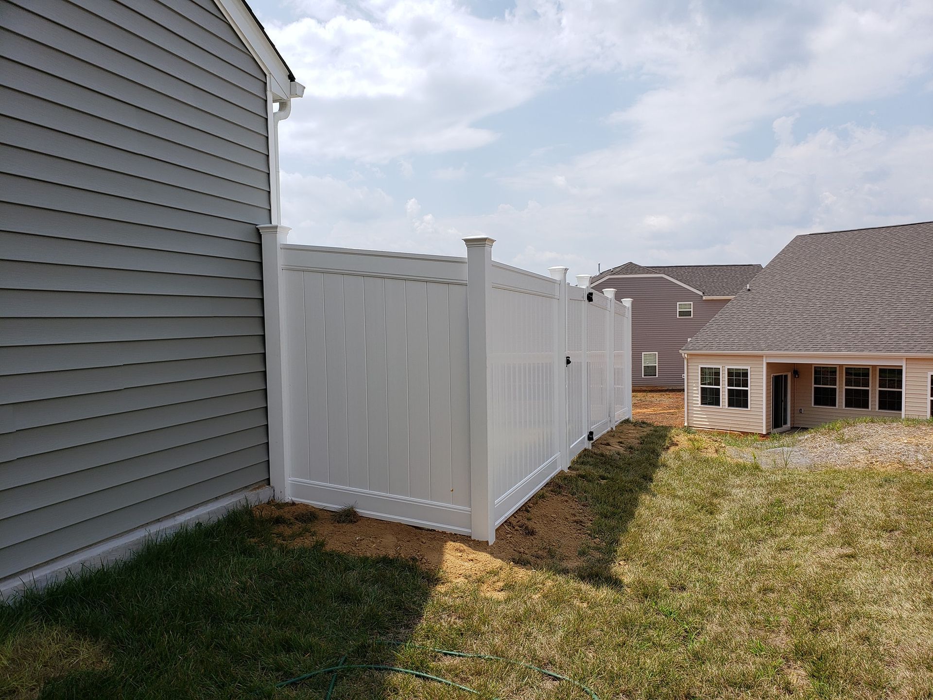 Backyard beside gray house and white vinyl fence, with lawn and neighboring homes under a cloudy sky