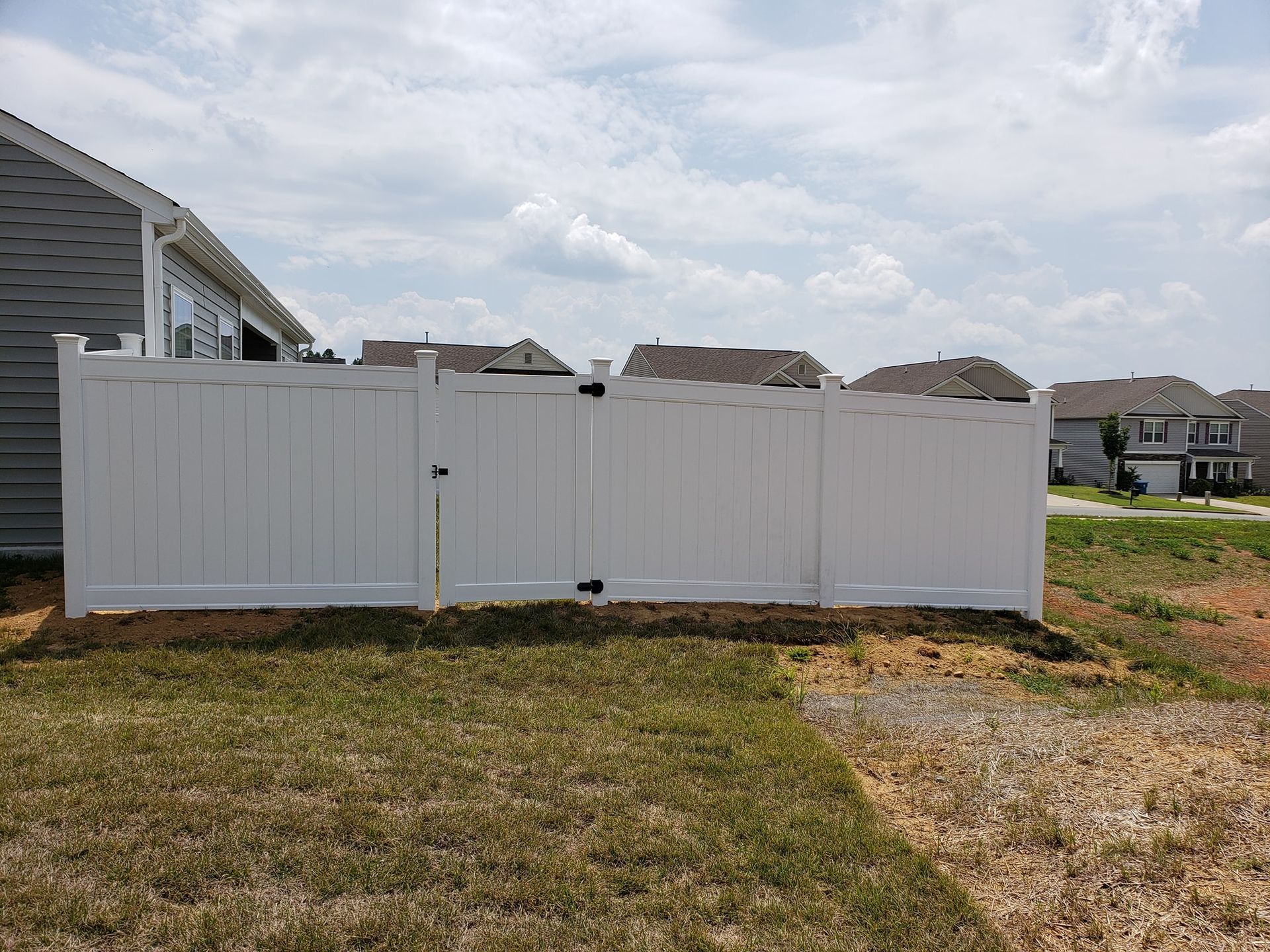 White vinyl fence beside a house in a grassy yard, with roofs and sky visible behind it.