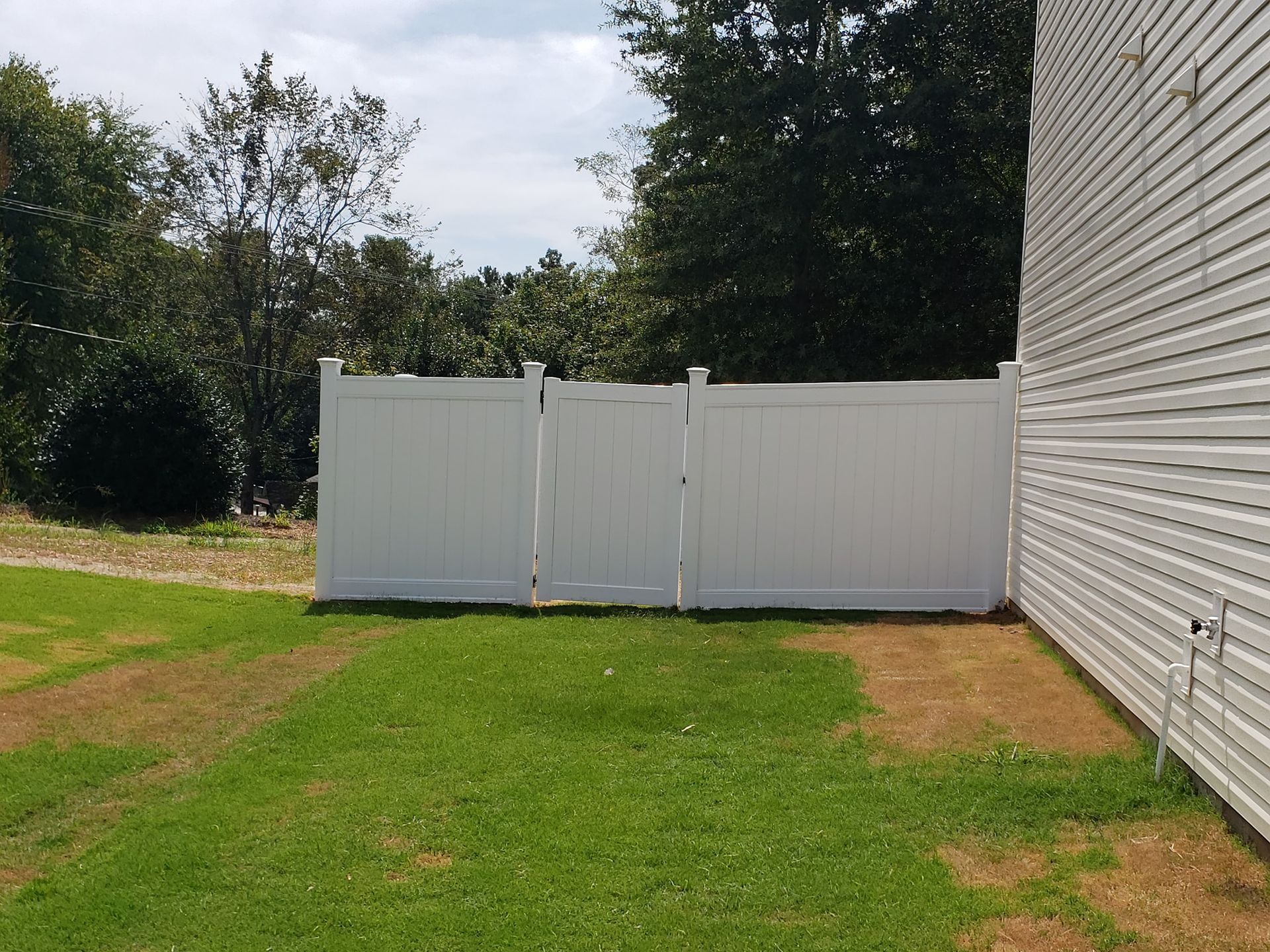 White vinyl fence beside a house, bordering a green lawn with trees in the background.