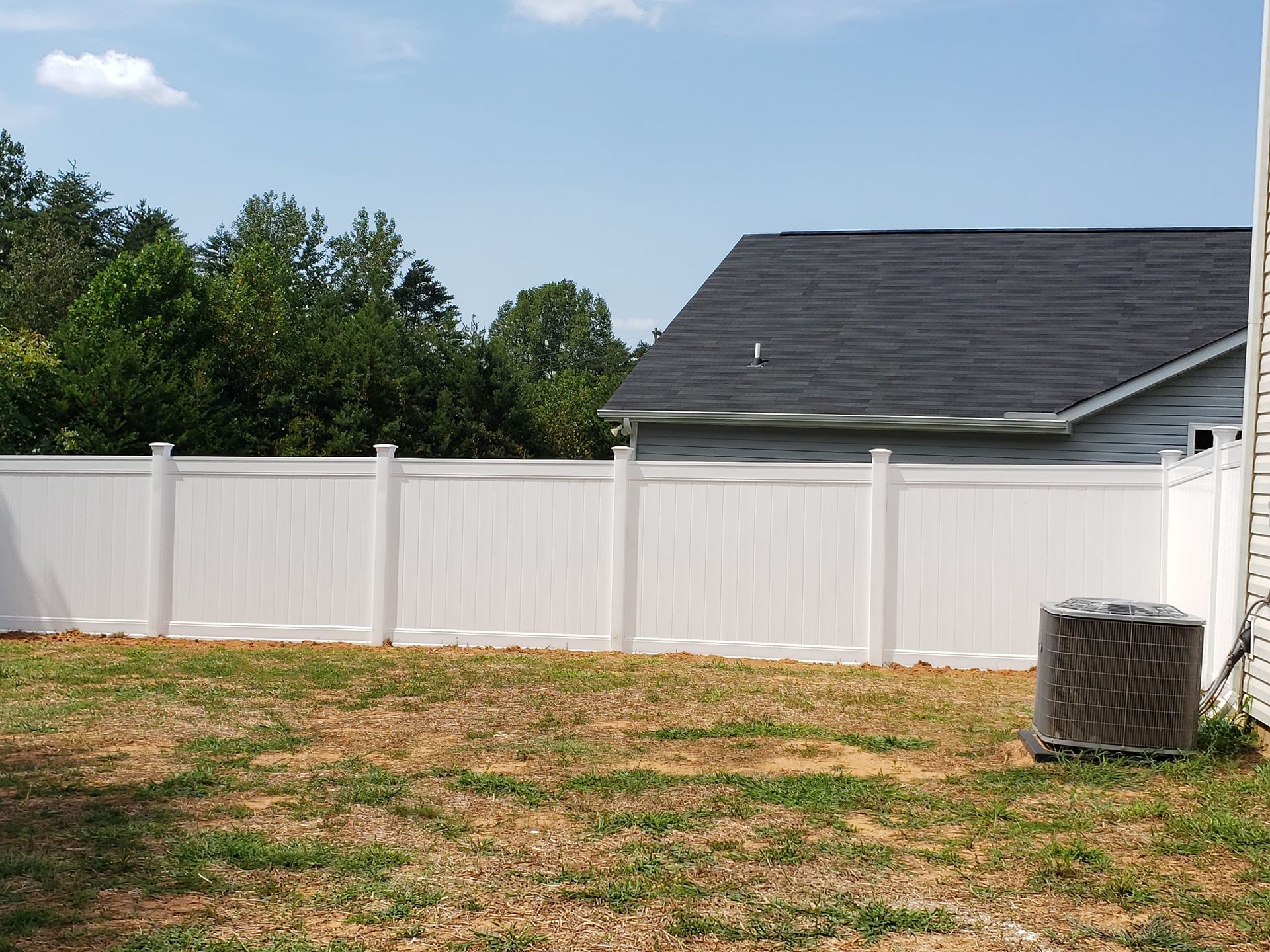 Backyard with white vinyl fence, grassy yard, trees, and a house roof under a blue sky