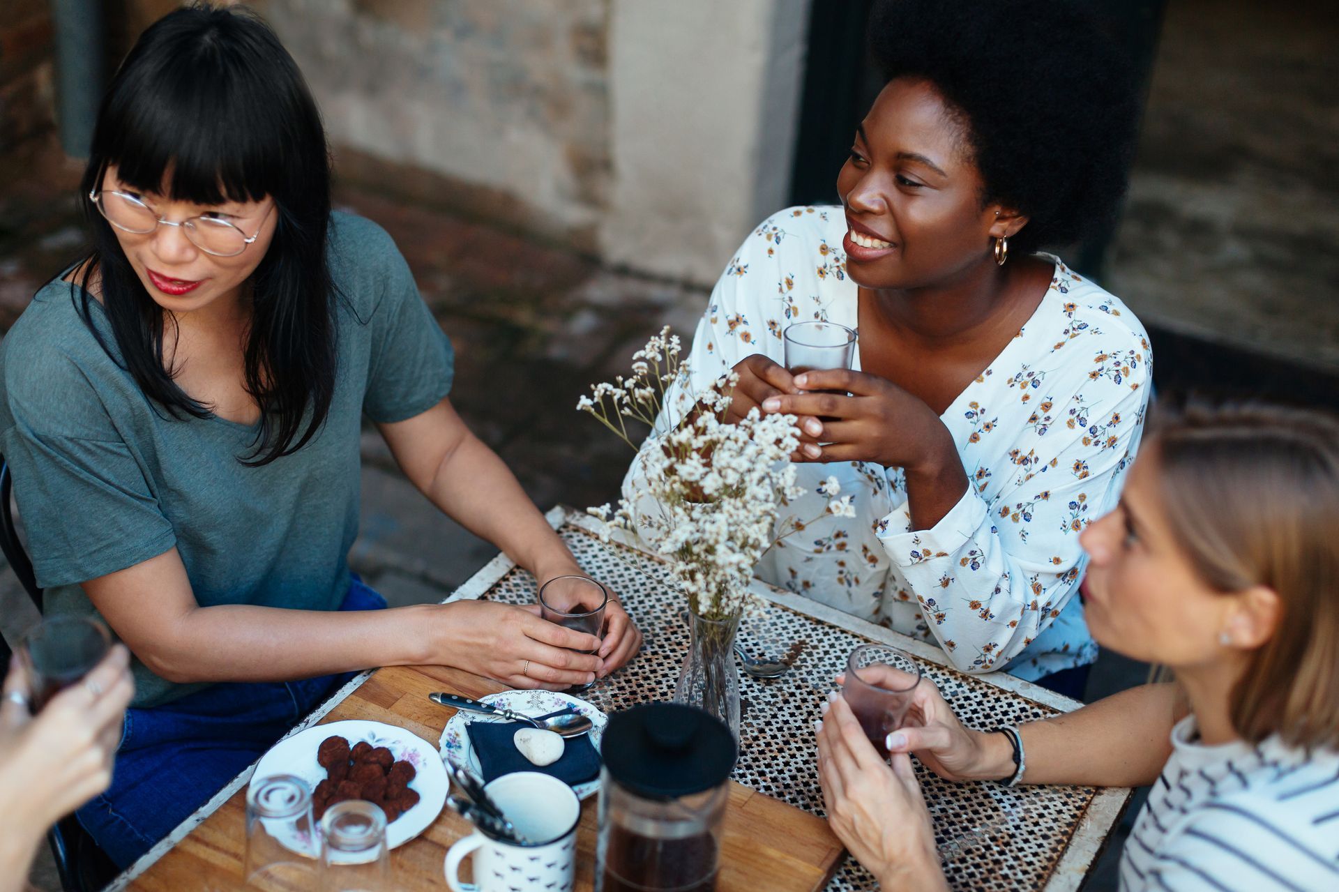 Three women sit at an outdoor table, smiling and chatting, with a small vase of flowers.
