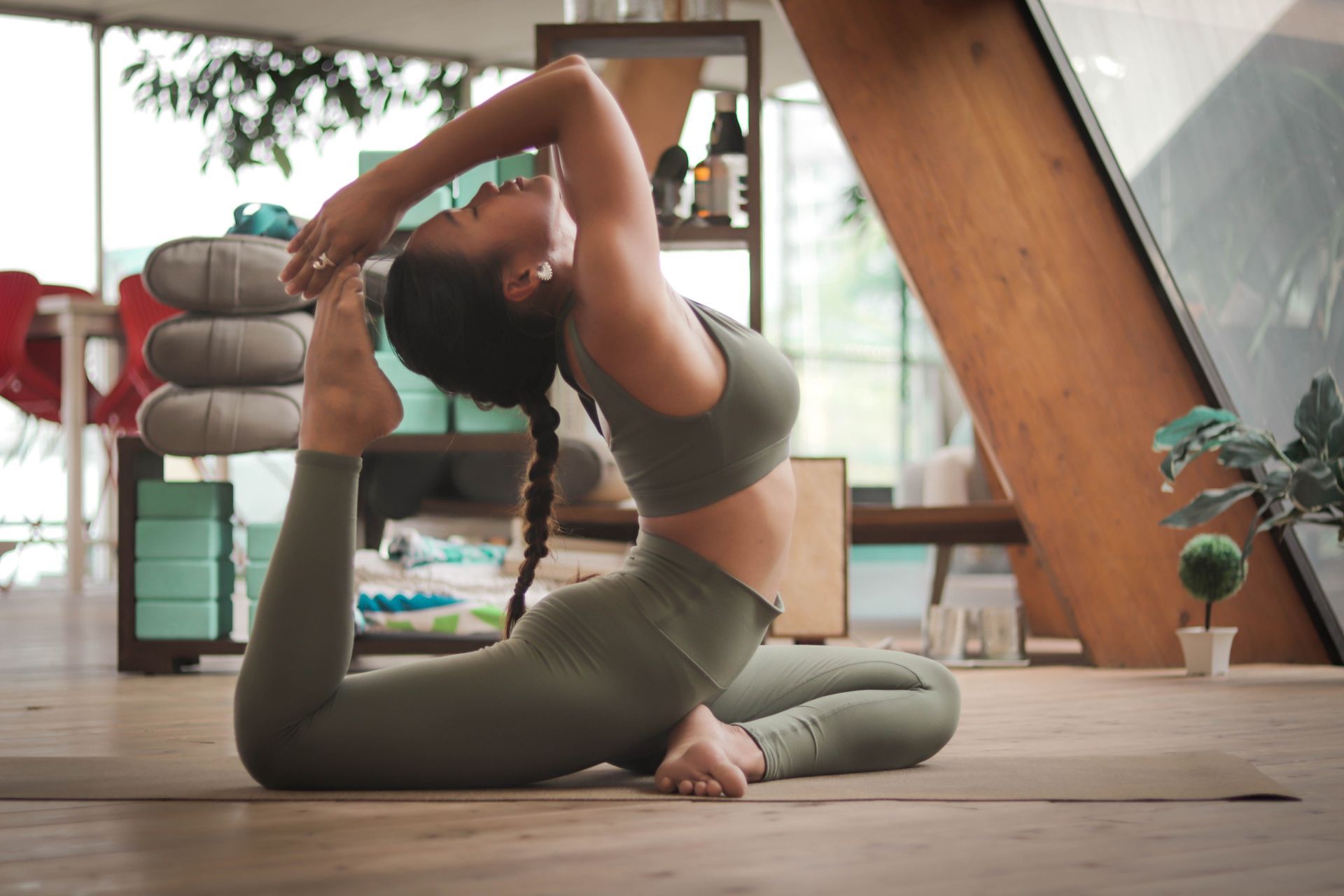 Woman in olive activewear, performing yoga pose, reaching for foot. Indoors, natural light.