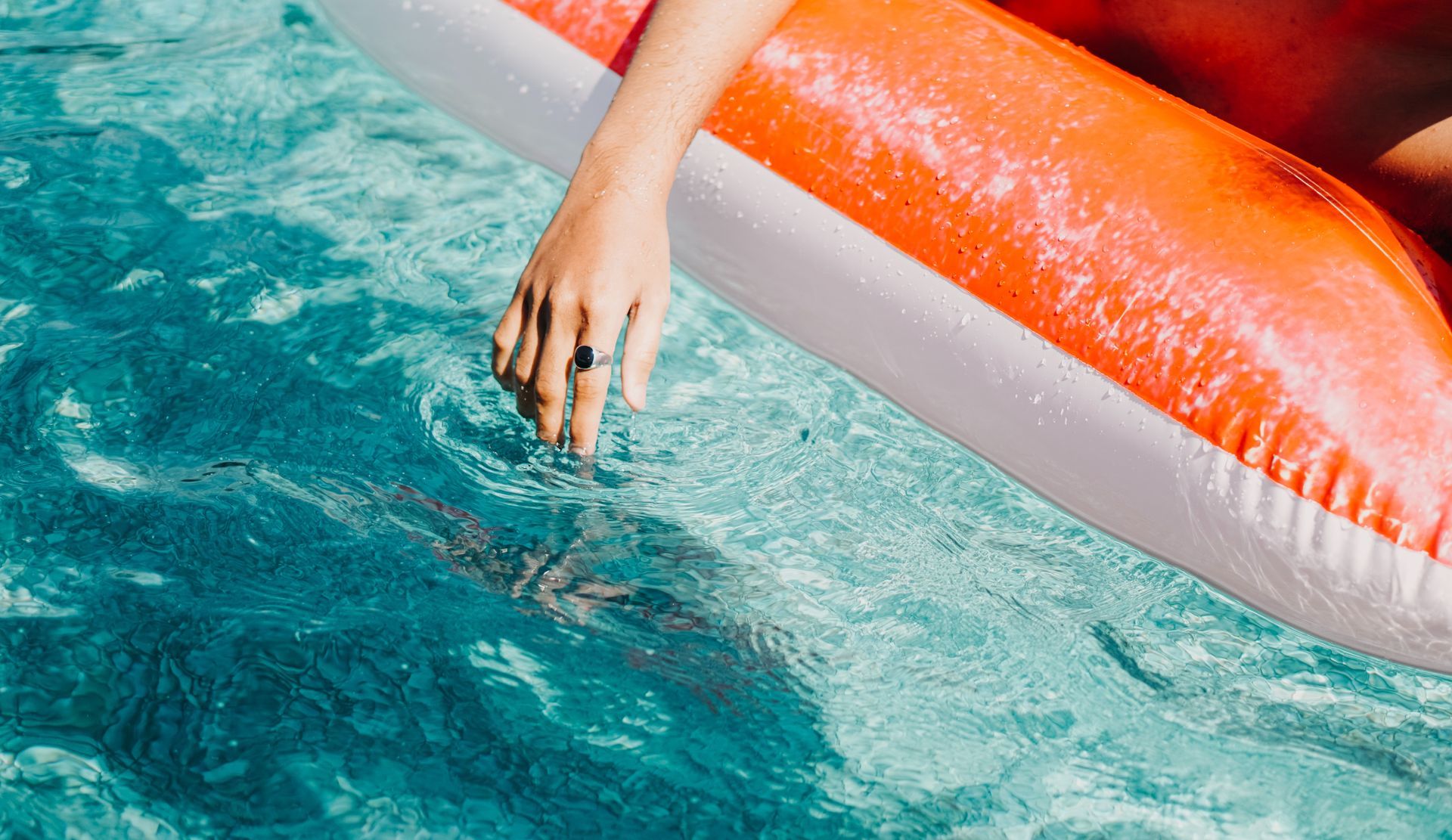 Hand with a ring dips into a pool of turquoise water while resting on a red and white pool float.