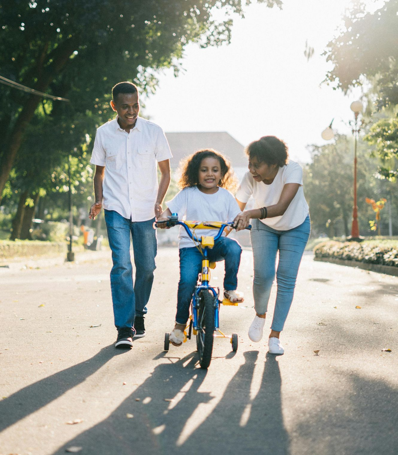 Family helps a child ride a bike on a sunny park path.