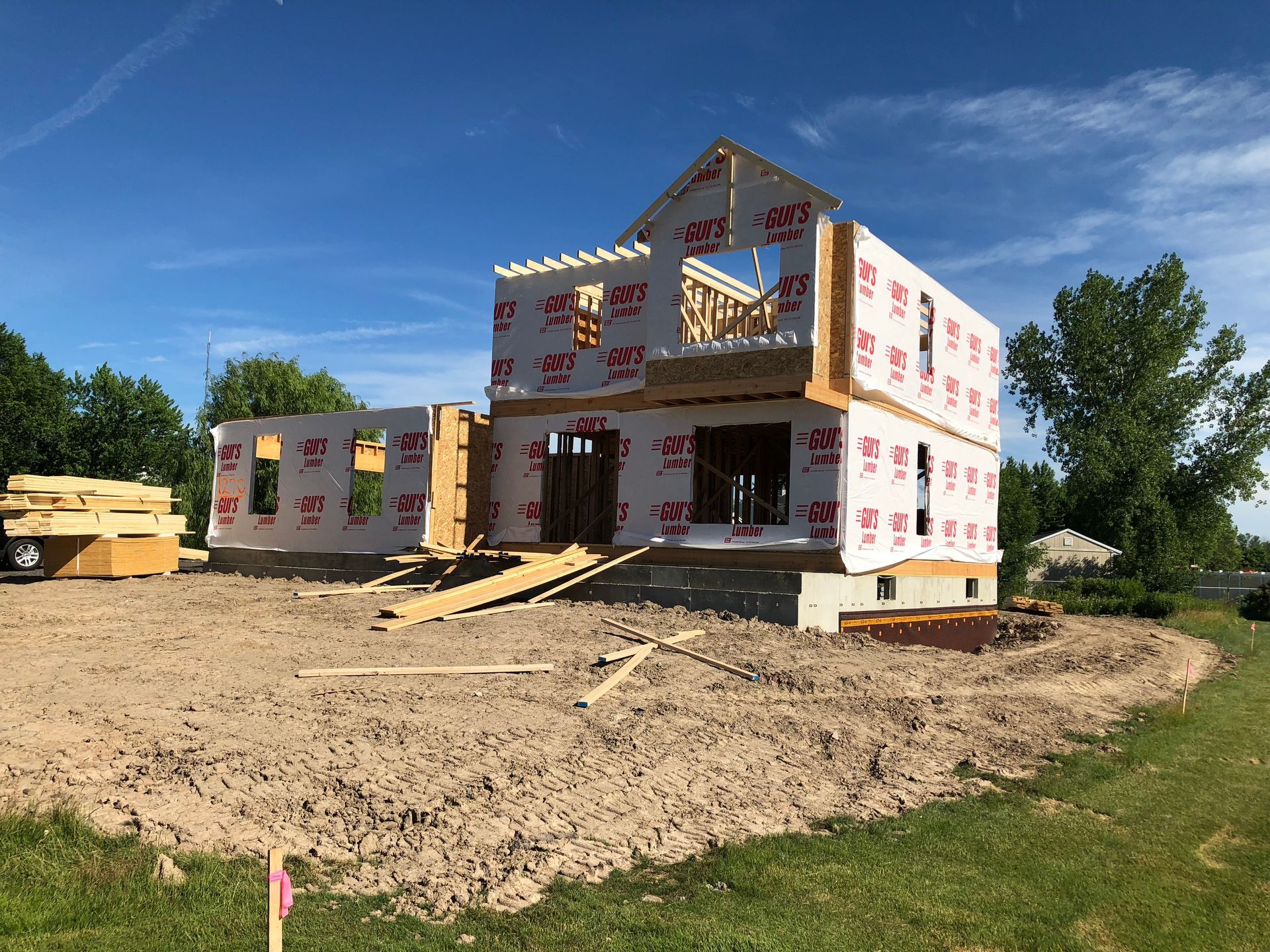 Two-story house under construction, framed with wood against a blue sky, surrounded by trees.