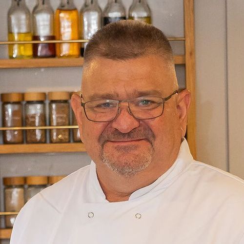 Paul wearing a chef 's coat is standing in front of a spice rack.