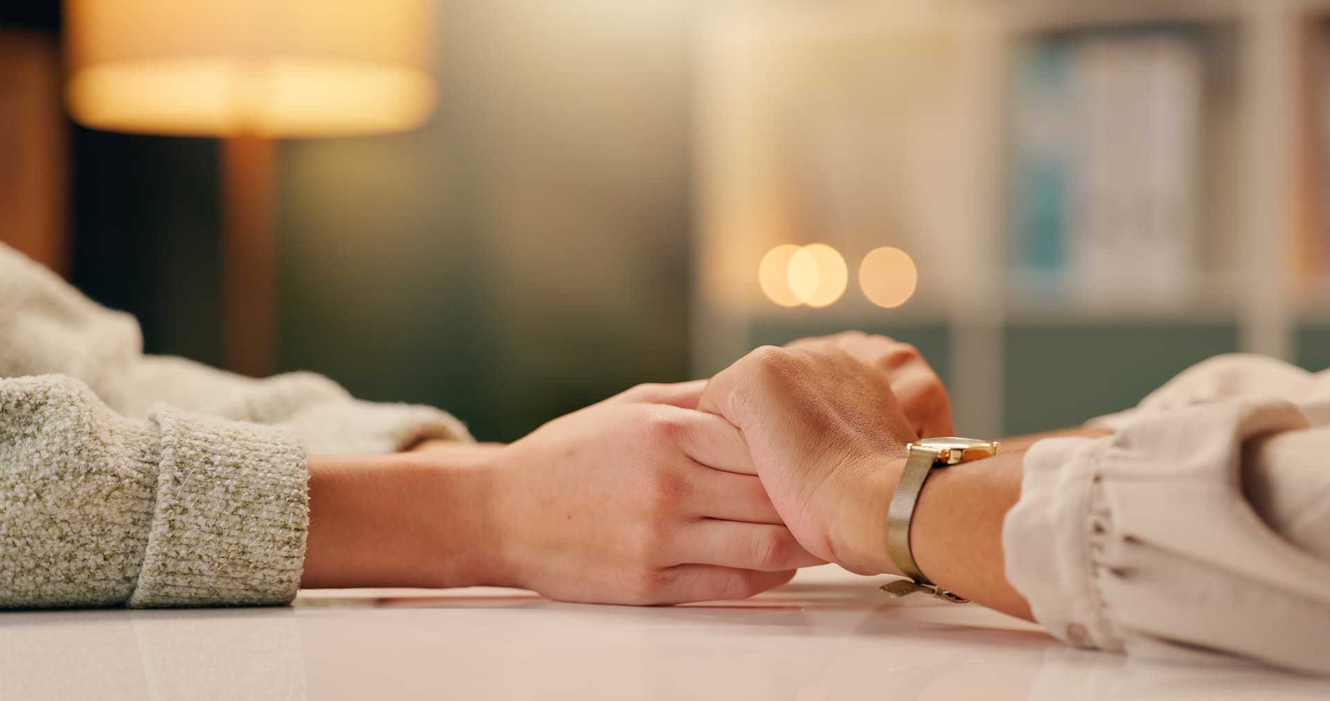 Two people hold hands across a table in a softly lit indoor setting.