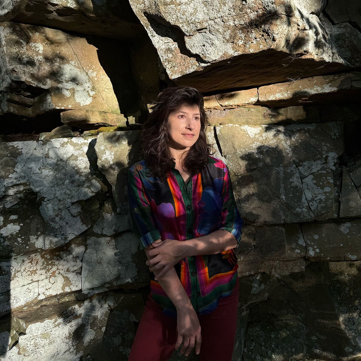 Picture of Cristina standing against a wall of rock outside with the sun shining on their face. 