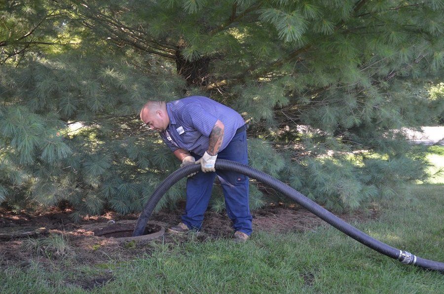Man in blue uniform, emptying septic tank, holding black hose, green grass and trees in background.