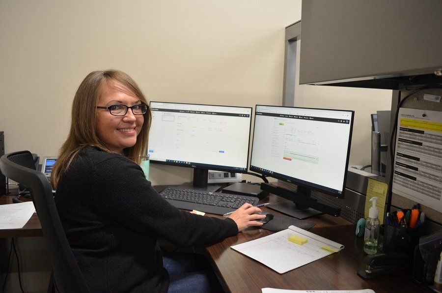 Woman smiling at desk with dual monitors, typing and using a mouse.