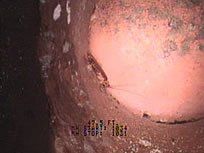 Inside a rusty pipe, a camera view shows a large, pinkish obstruction.