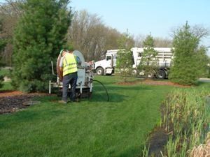 Man operating equipment, likely spraying, near a truck on a grassy lawn with trees and a small body of water.