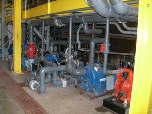 Industrial equipment: pipes, pumps, and valves. Gray and red components on a concrete floor, beneath yellow beams.