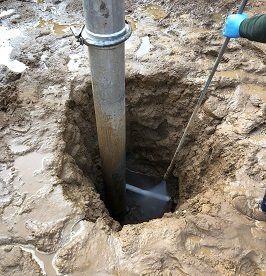 Person using a tool to clean a metal pipe in a muddy hole outdoors.