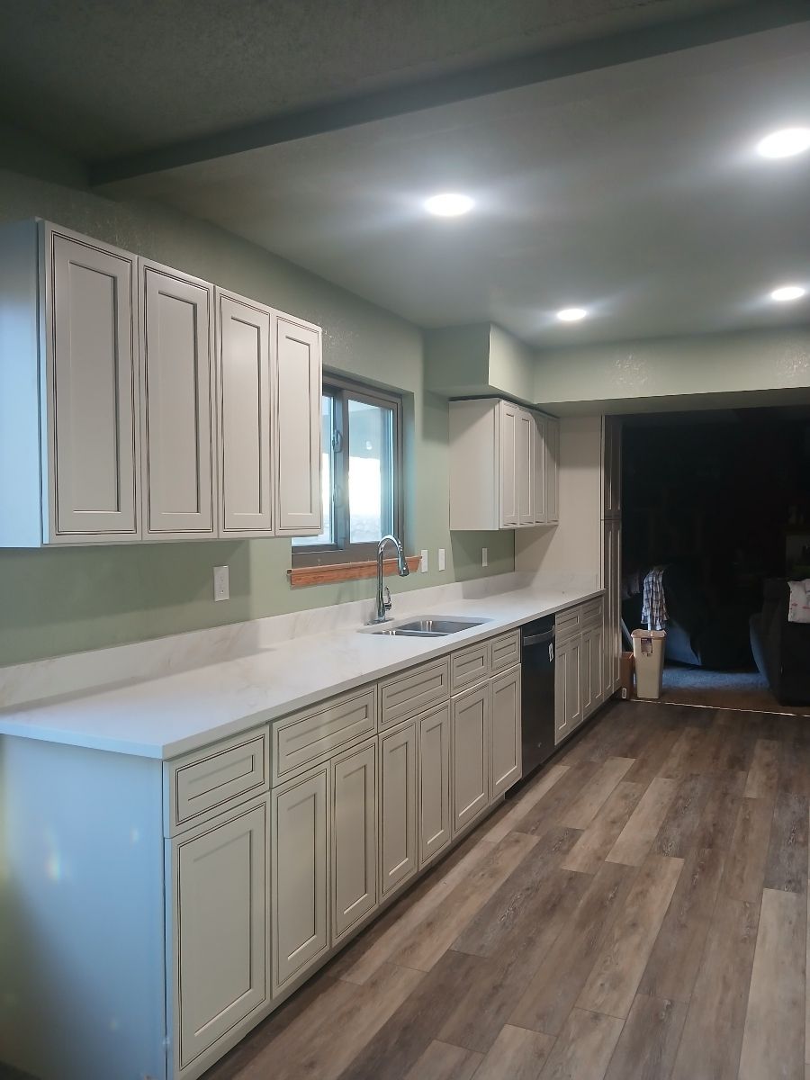 Modern kitchen with white cabinets, light countertops, sage green walls, and wood-look flooring under recessed lighting.