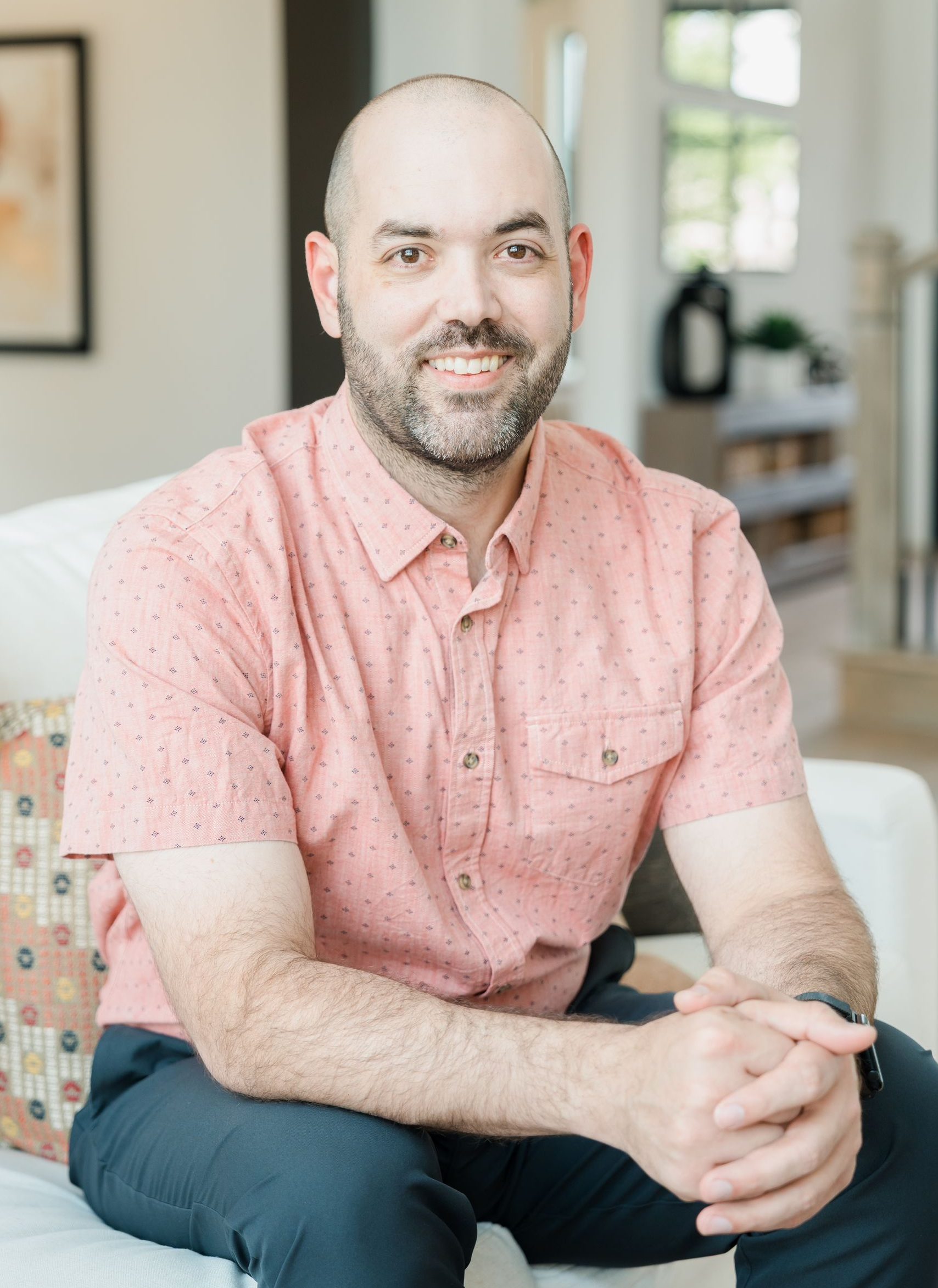 A man in a suit and white shirt is sitting on a couch and smiling for the camera.