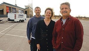 Three people standing in a parking lot, a building and trailer in background.