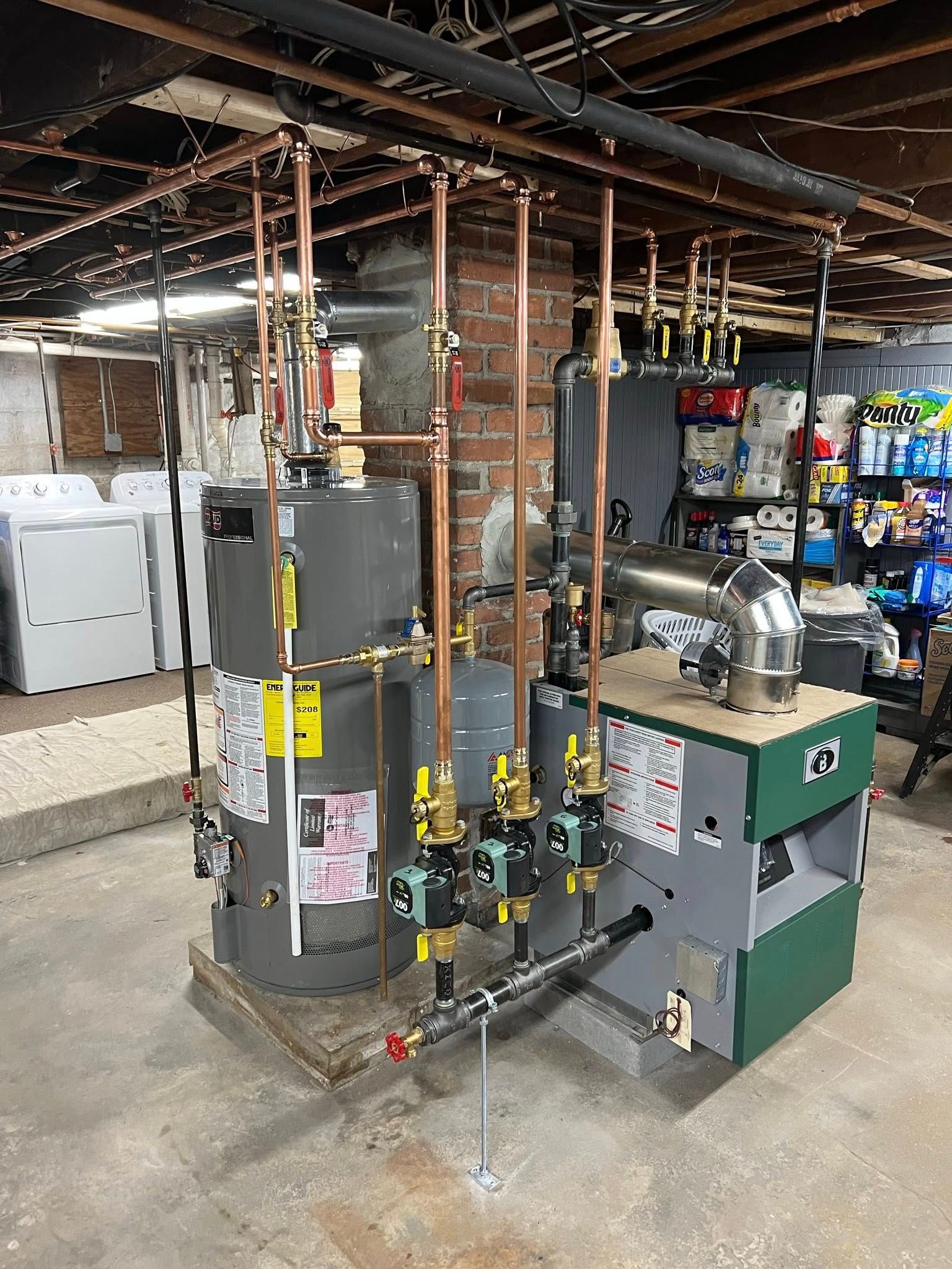 A basement utility room featuring a grey water heater and a green boiler connected by copper piping and circulator pumps.