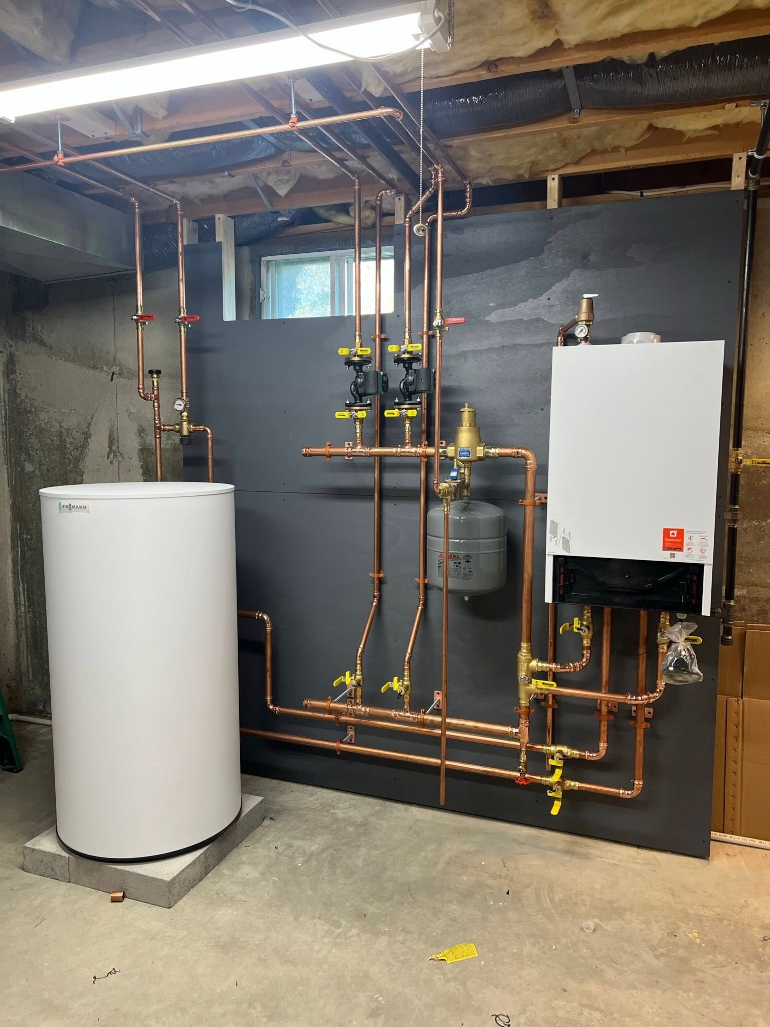 A white boiler and matching water tank connected by copper piping against a dark wall in a basement mechanical room.