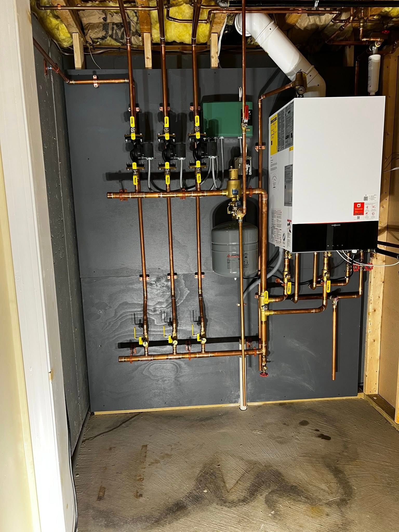 A white wall-mounted boiler unit connected to a complex copper pipe system with valves against a grey wall in a basement.