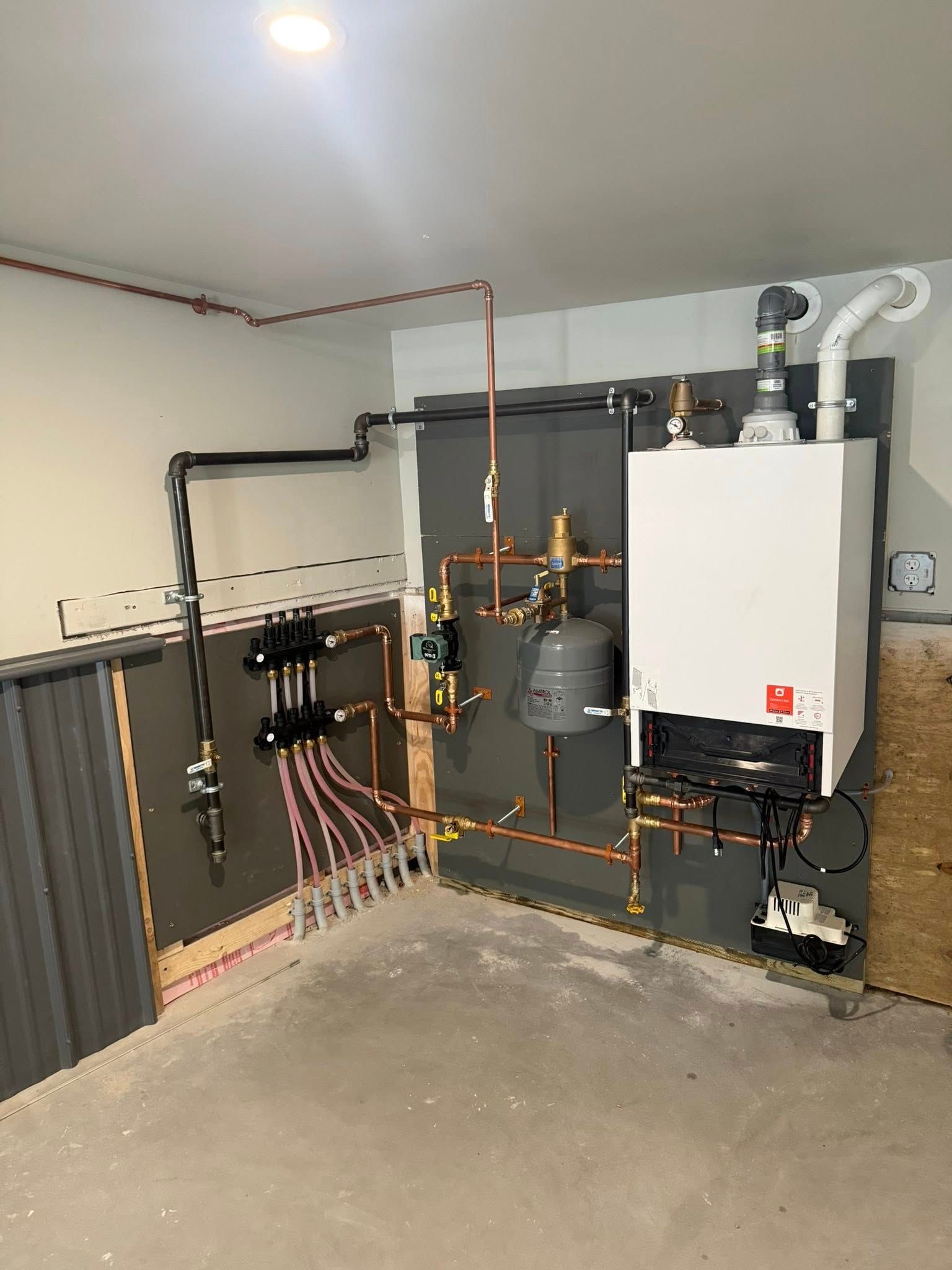 A wall-mounted boiler and water distribution manifold system with exposed copper pipes against a grey and wooden wall.