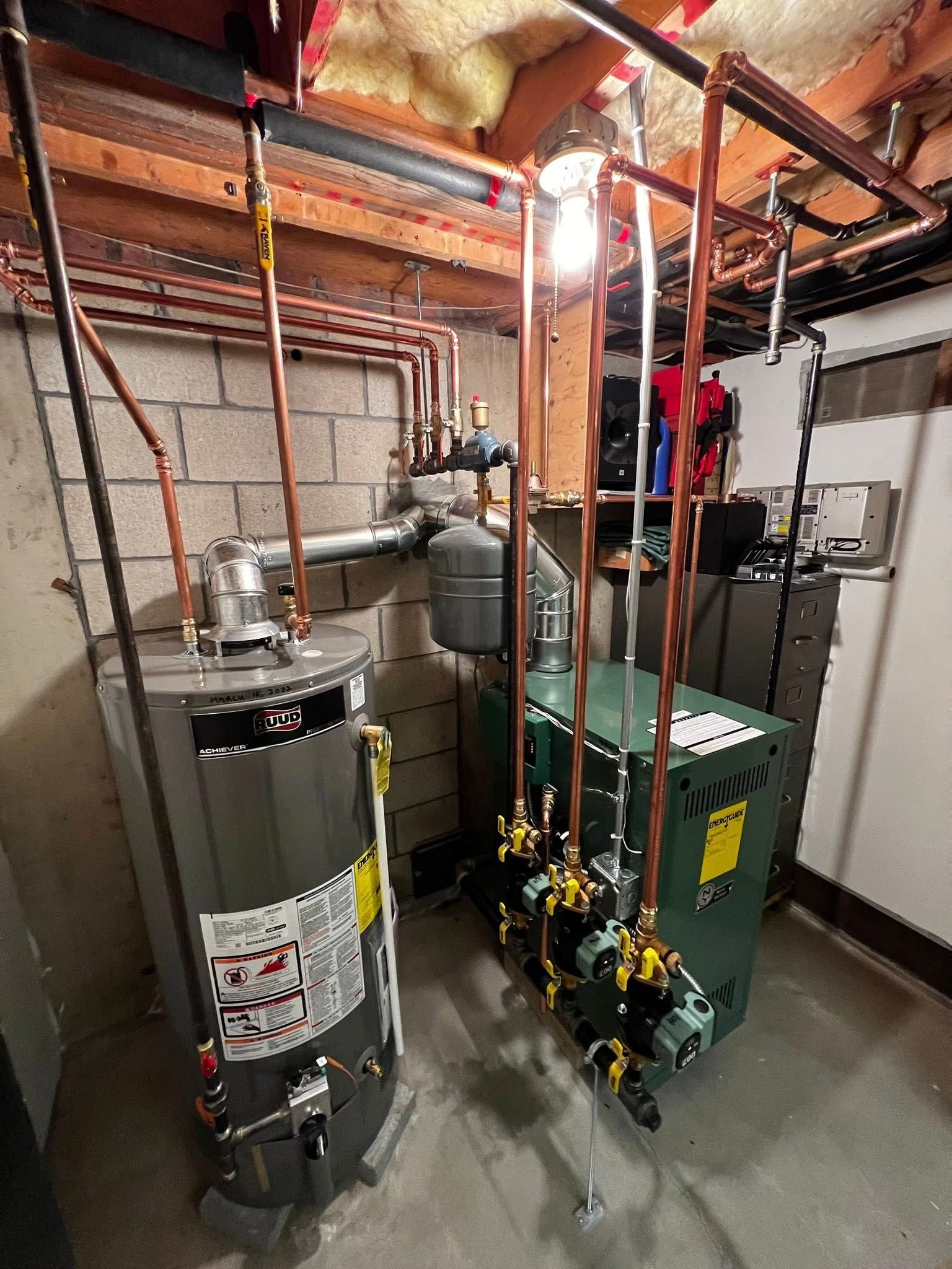 A water heater and a green boiler with copper pipes are installed in a utility room with cinder block walls.