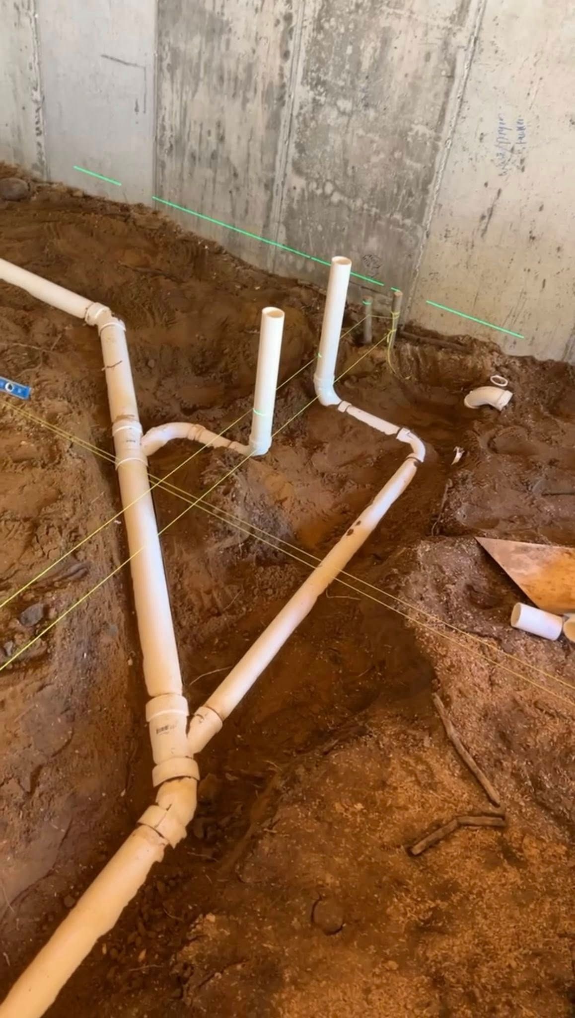 White PVC plumbing pipes installed in a dirt trench next to a concrete wall at a construction site.