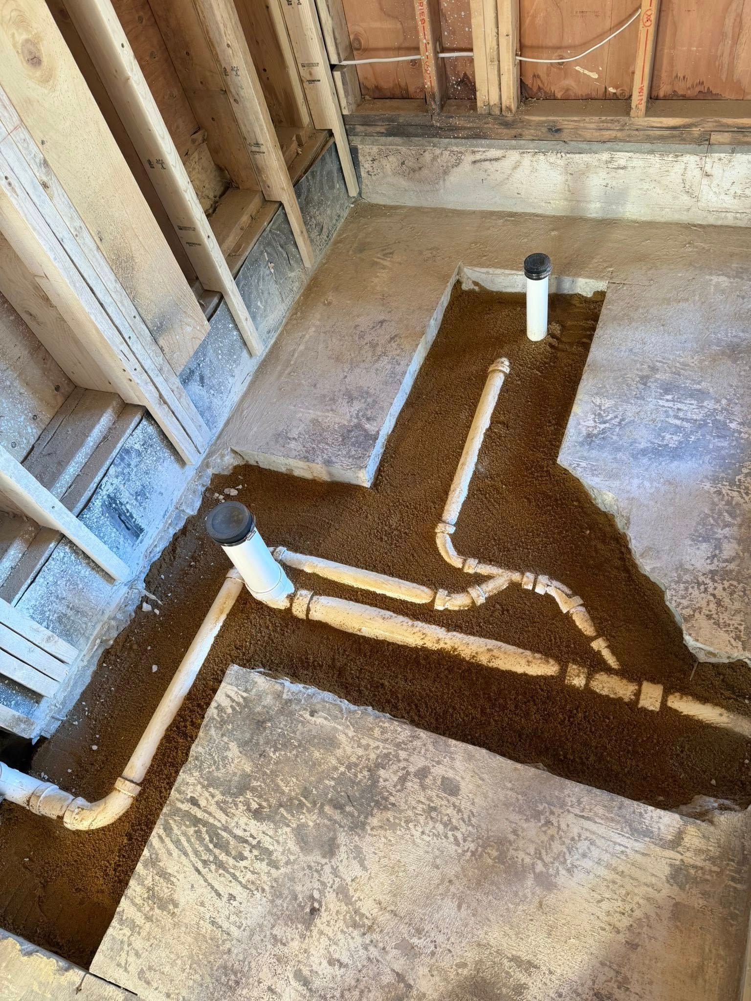 Plumbing pipes installed in a trench dug into the dirt floor of a room under construction with exposed wall studs.