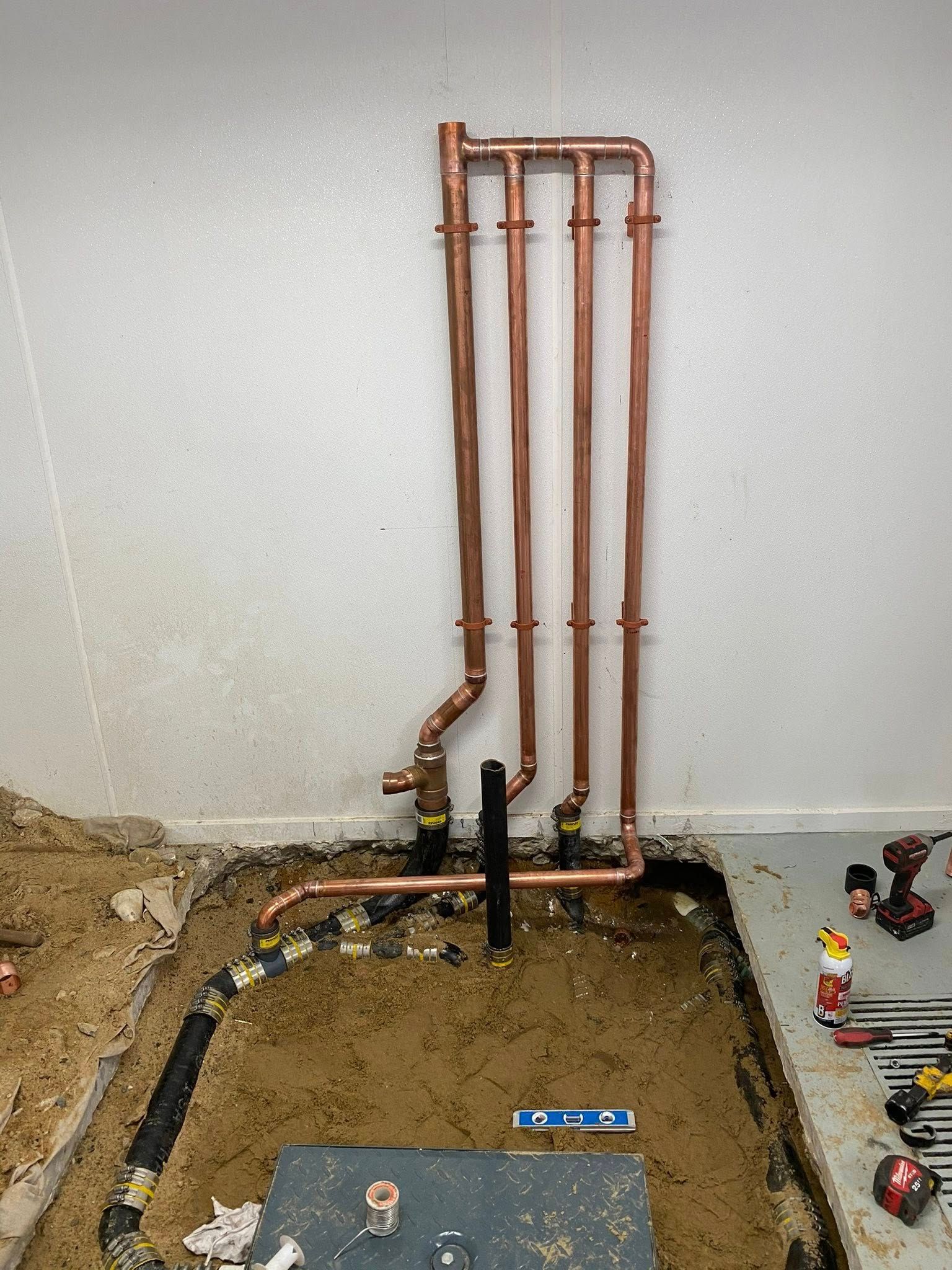 Copper piping installation against a white wall with pipes leading into a dirt-filled cutout in a concrete floor.