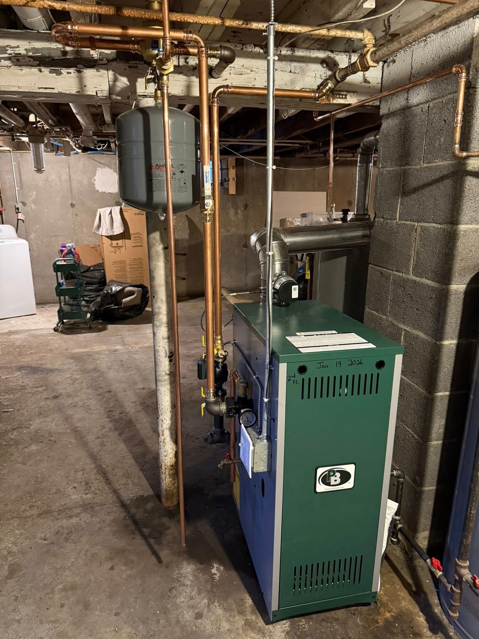 A green residential boiler and copper plumbing pipes in a basement with concrete walls and a dirt floor.
