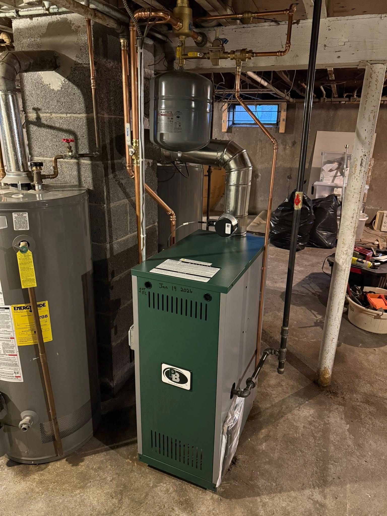 A green and grey boiler stands beside a hot water tank in a basement with exposed pipes, a vent, and a concrete floor.