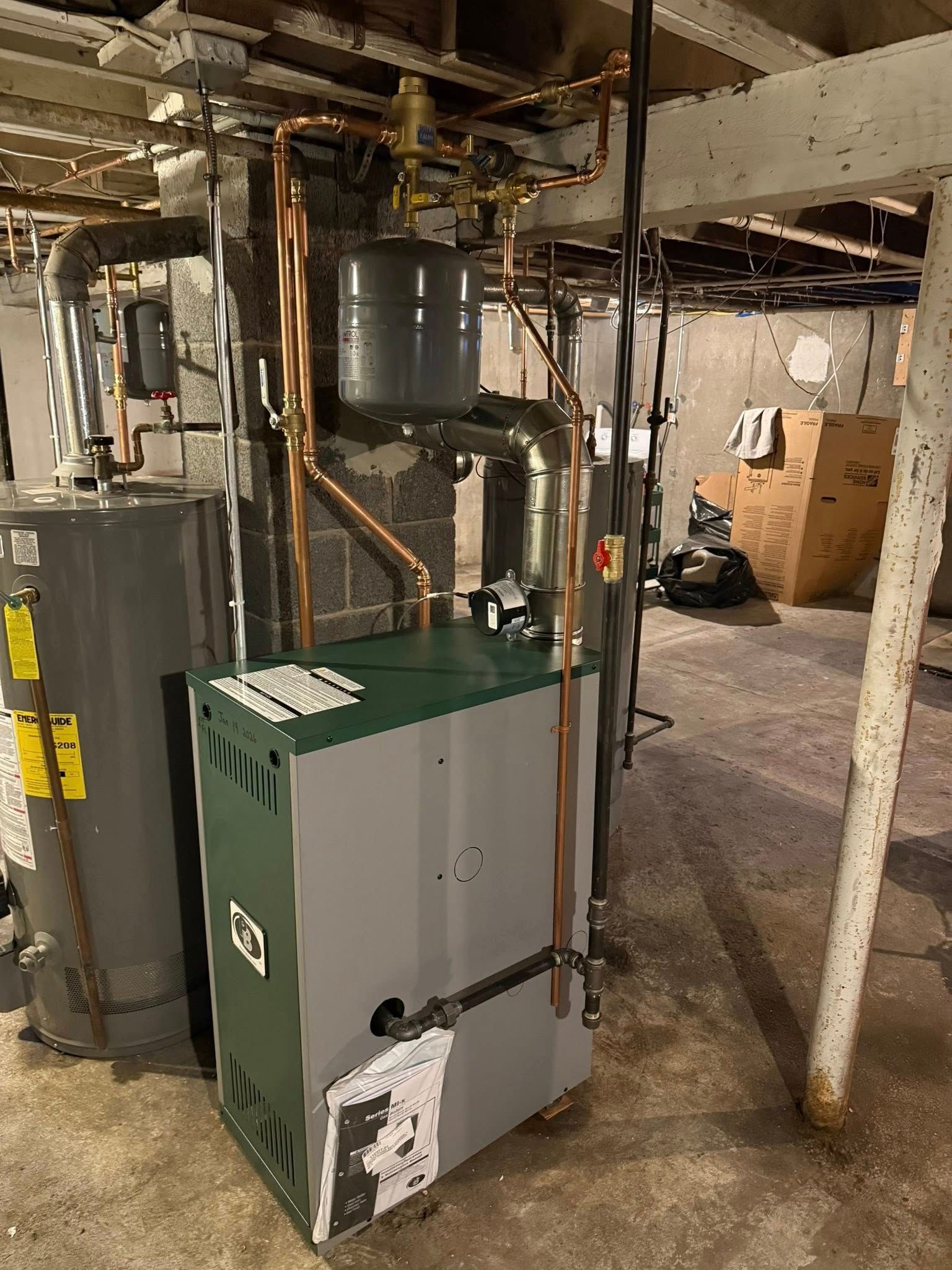 A grey and green furnace with copper pipes and a small expansion tank, located in a basement utility area.