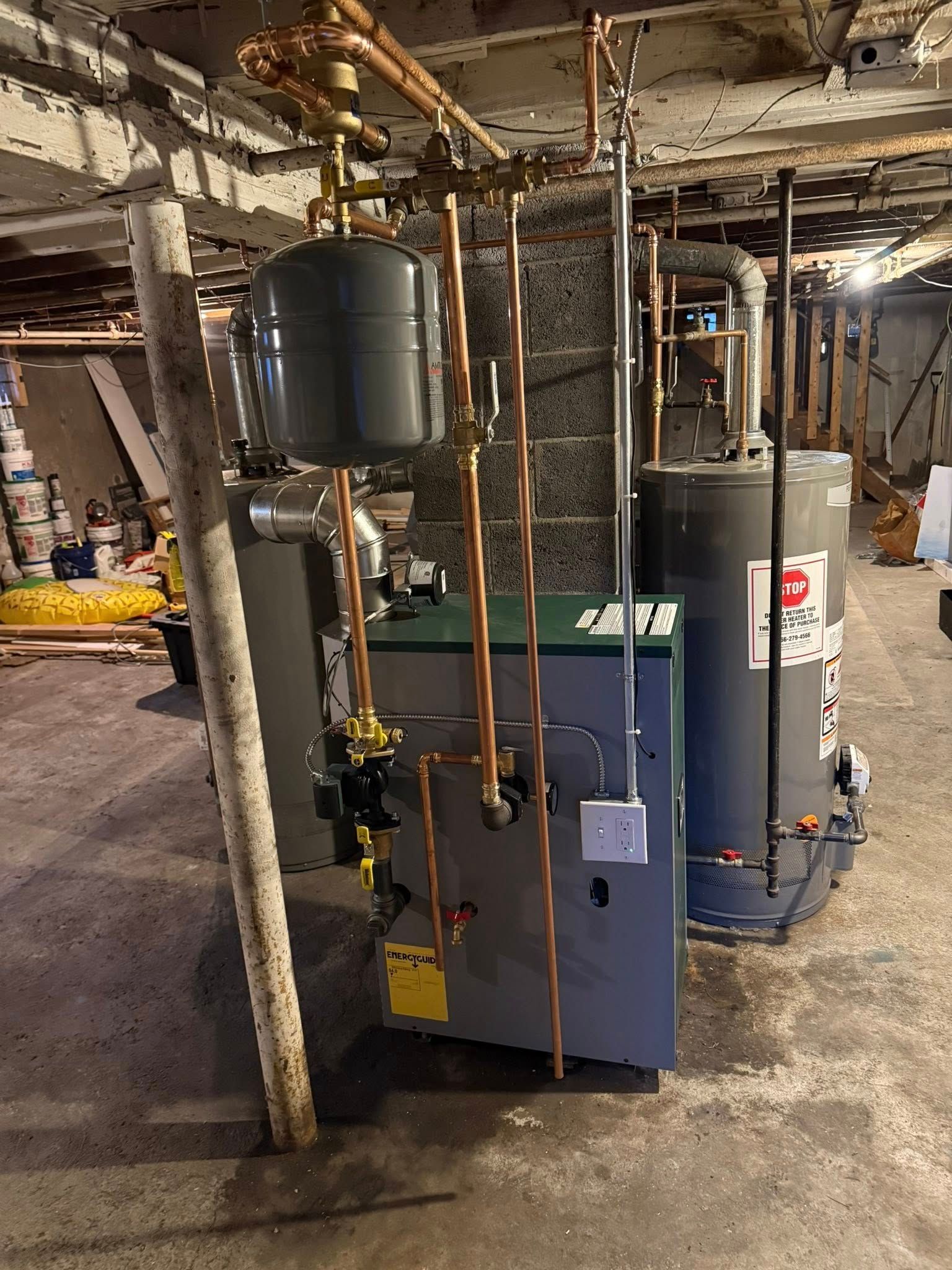 A basement view of a green residential boiler unit with copper plumbing, an expansion tank, and a grey water heater.