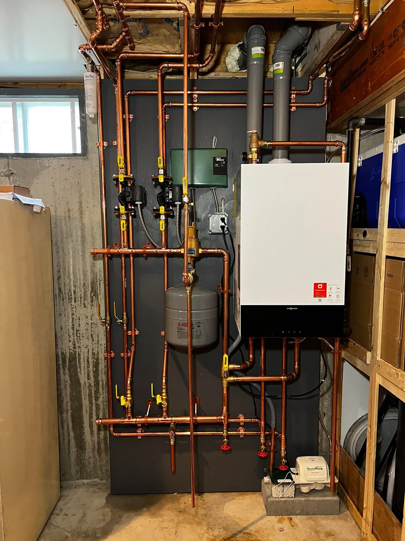 Wall-mounted boiler unit with complex copper plumbing and pipes, mounted against a grey panel in a basement.
