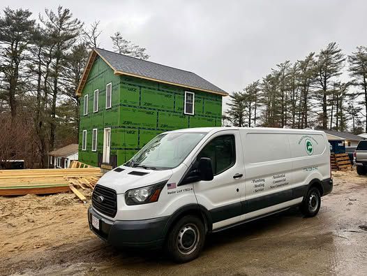 A white utility van parked in front of a partially constructed green residential building on a gravel lot.