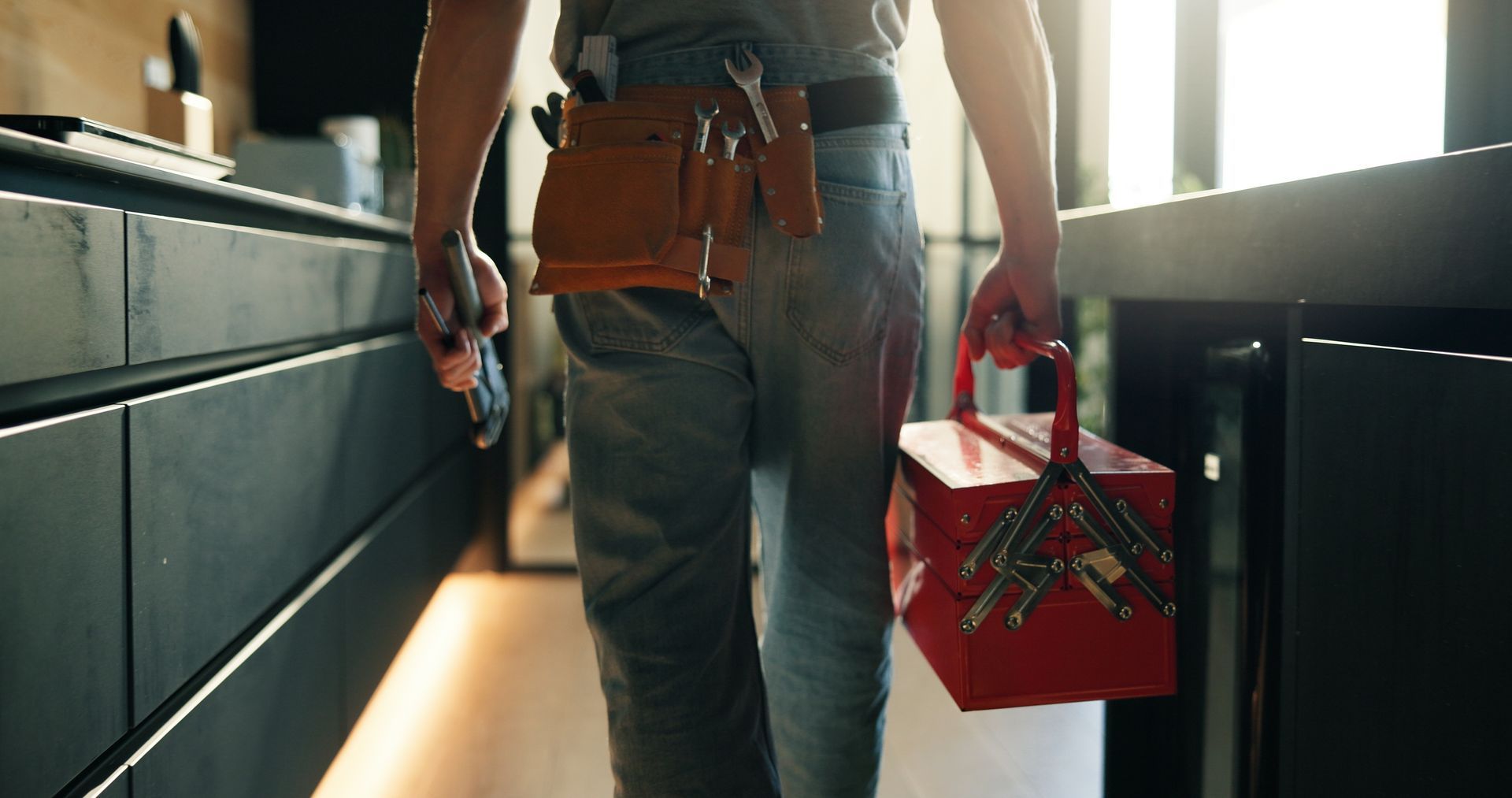 A worker wearing a tool belt walks through a kitchen carrying a red metal toolbox.
