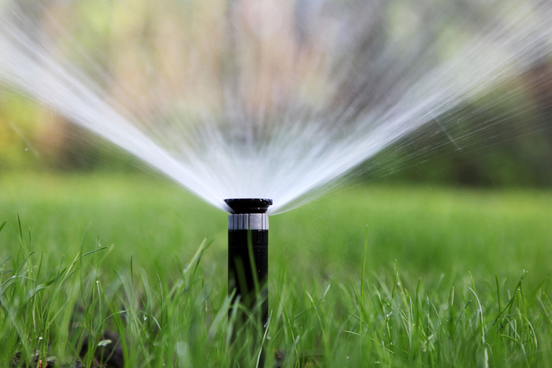 A black irrigation sprinkler head spraying a wide arc of water over a lush, green grassy lawn.