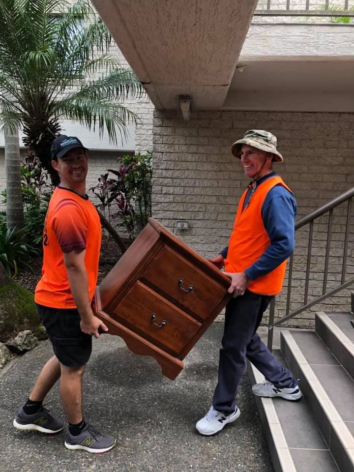 Two Men In Orange Vests Carry A Wooden Nightstand Up Outdoor Stairs. One Smiles