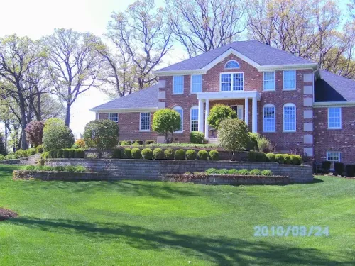 A large two-story brick house with a multi-level retaining wall and manicured landscaping under a bright blue sky.