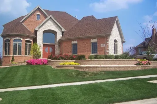 A two-story brick and stone house with a brown roof and landscaped front yard under a blue sky.