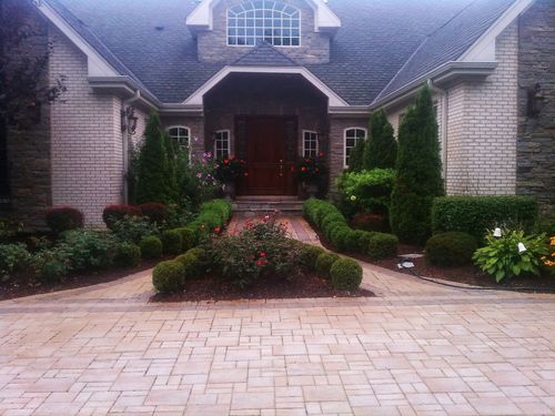A paved driveway leads to the front entrance of a stone house with symmetrical landscaping and evergreen shrubs.