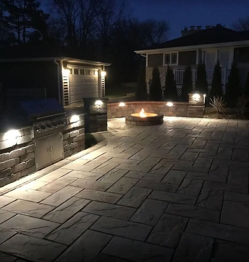 A stone patio at dusk with a lit fire pit, an outdoor grill station, and ambient landscape lighting.