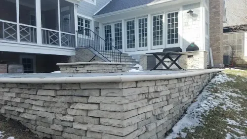 A stone patio with a built-in fire pit and retaining wall, set against a house with a screened porch and large windows.